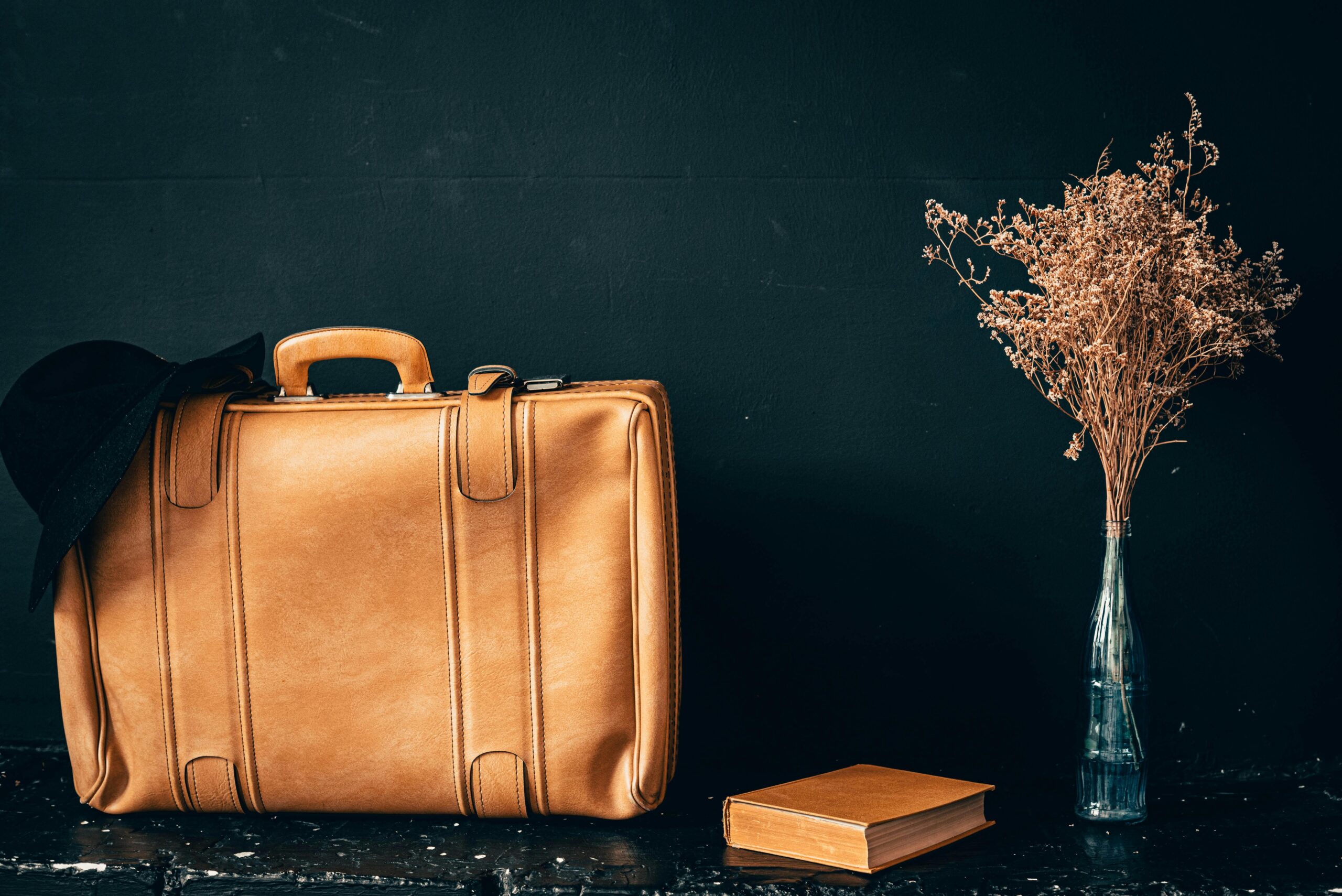 A vintage leather suitcase paired with dried flowers in a vase and a book on a dark background.