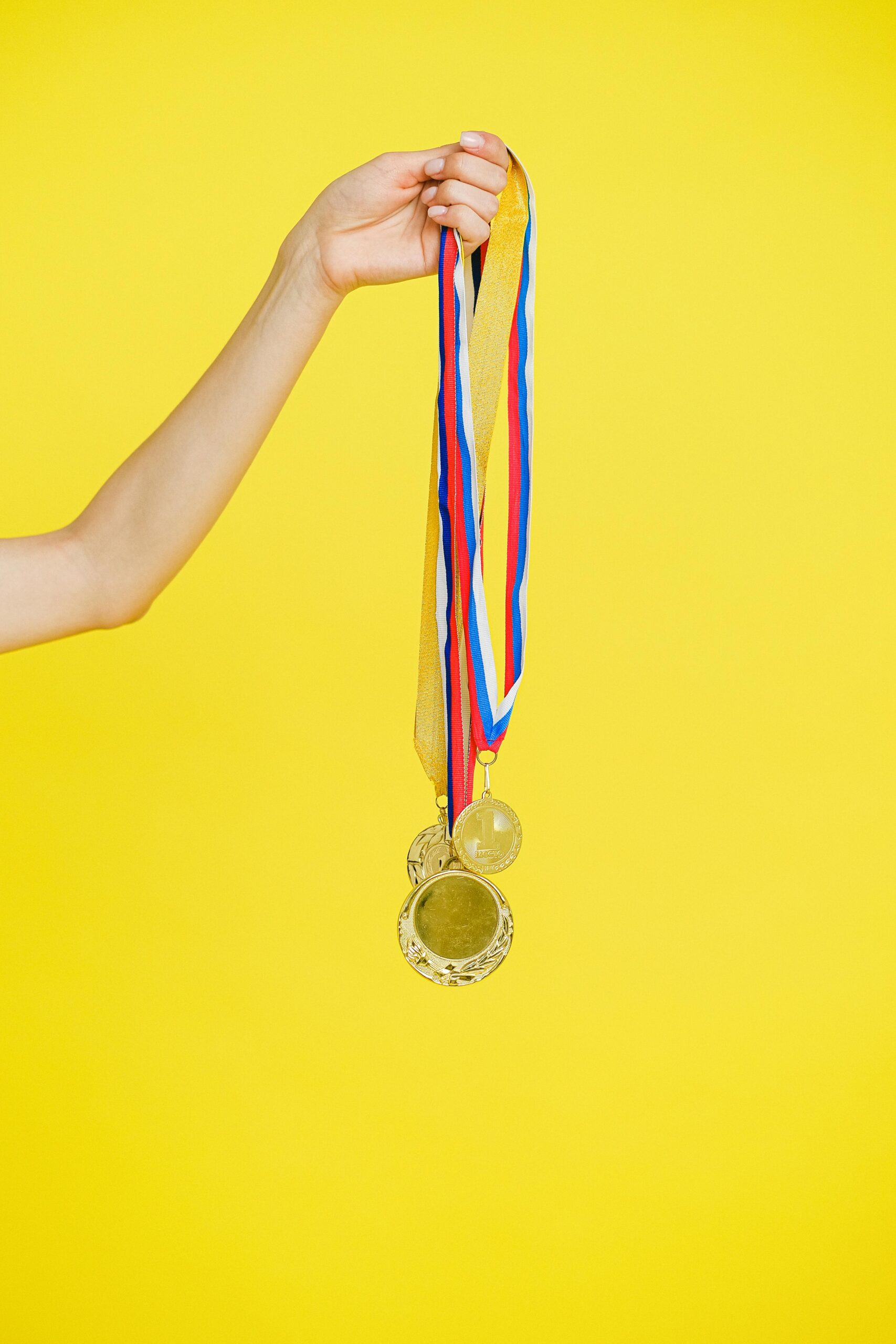 A hand holding multiple gold medals with colorful ribbons on a vibrant yellow background.
