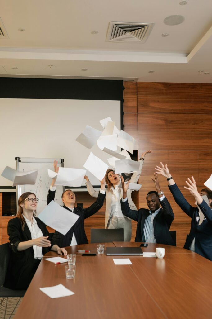 A diverse office team joyfully throws papers in a modern meeting room, celebrating success.